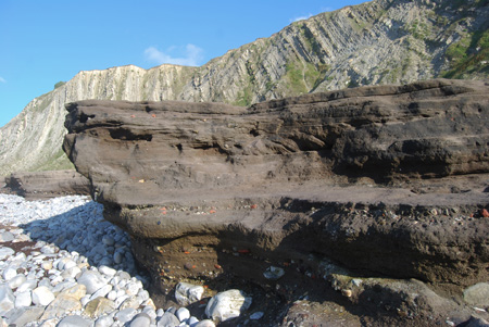 Vista en detalle de la playa cementada de Gorrondatxe (Getxo-Sopela) cuyos materiales de color oscuro se disponen discordantes sobre los estratos claros del acantilado e incluyen diversos tecnofósiles como restos de ladrillos, plásticos, etc. El espesor estimado de estos depósitos antropocenos es de unos 1,60 m.